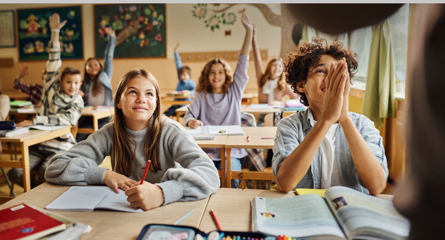 Students seated around desks, some with hands raised, classroom walls show educational materials.