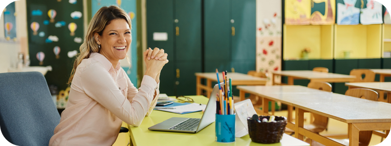Teacher smiling at the camera, seated at a desk at the front of a classroom