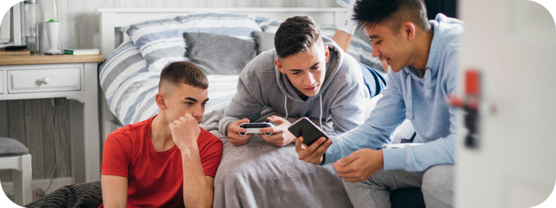 A group of young men gathered in a bedroom, looking at a mobile; one is holding a game console.
