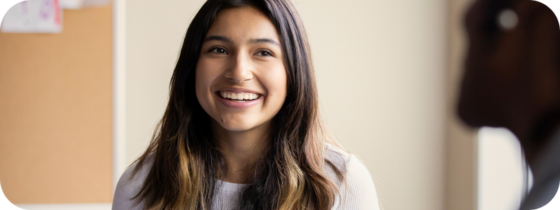 A young woman with a bright smile standing indoors against a wall.