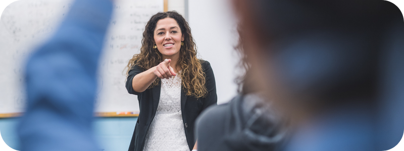 A female teacher standing at the front of a classroom, pointing at a student with raised hand.