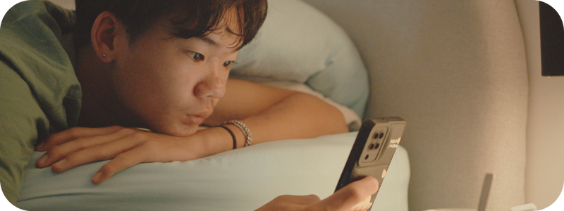 A young male is lying comfortably on a bed, their face partially visible looking at their mobile.