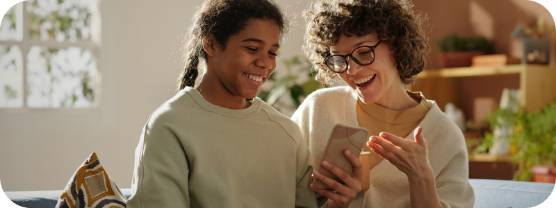 Woman with glasses and teen with braids smile while looking at a smartphone indoors.