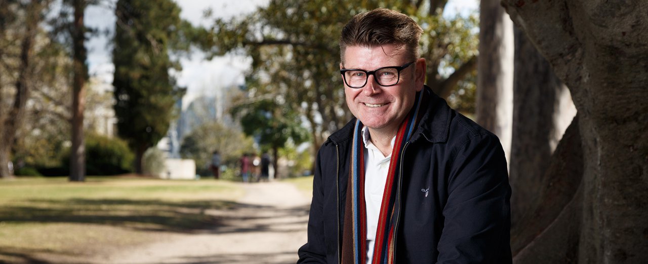 Photo of a smiling middle-aged man with short brown hair, wearing glasses, a white polo shirt, navy jacket and red scarf, sitting against a tree, parklands behind.