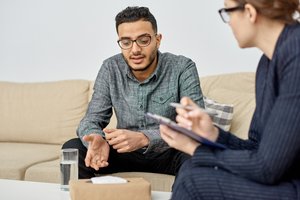 A man and woman are seated on a couch, engaged in a discussion. The woman is wearing glasses and holds a pen and notepad.