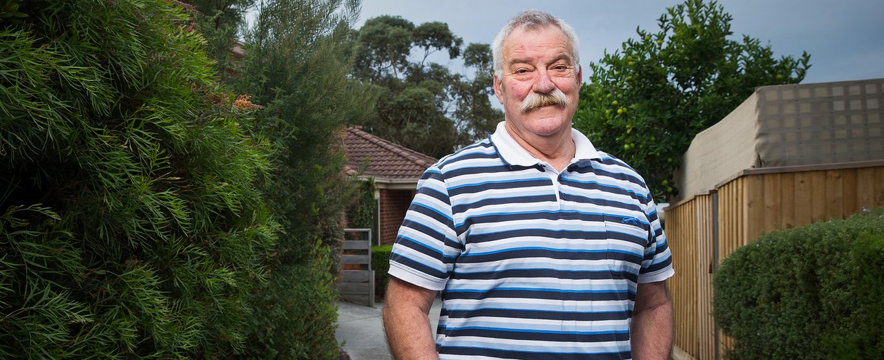 Photo of an older man with short grey hair and a grey moustache, wearing a blue and white striped polo shirt, standing in a suburban driveway, hands in pockets, smiling at the camera.