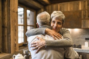 A woman with a gentle smile is hugging an older man indoors, in a kitchen setting. The background shows a wall and a window, adding warmth to the scene. Both individuals are dressed casually, conveying a sense of comfort and affection.