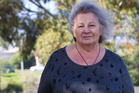 Woman in her 70s stands in front of a street and some trees, her face slightly in shadow as she smiles faintly for the camera.