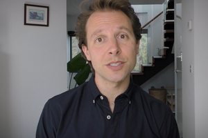 A male adult speaking to the camera, in an indoor setting with a bookcase and shelves in the background.