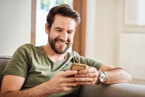 A young male sitting on a couch and looking at his mobile phone and smiling