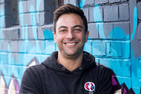 Headshot of Mario wearing a black hoodie in front of a graffiti wall, looking at the camera.