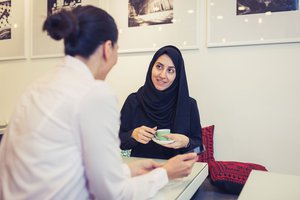 A woman wearing a hijab sits at a table, holding a cup of coffee, having a conversation with another individual